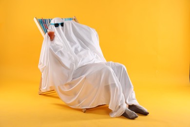 Photo of Person in ghost costume and sunglasses with glass of drink relaxing on deckchair against yellow background
