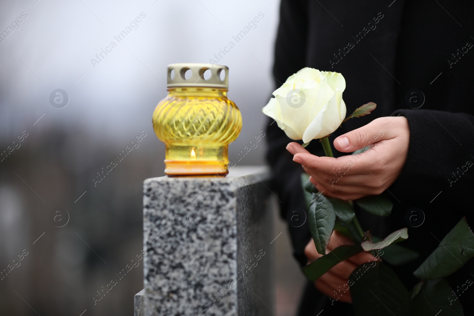 Photo of Woman holding white rose near grey granite tombstone with candle outdoors, closeup. Funeral ceremony
