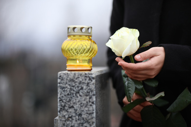 Woman holding white rose near grey granite tombstone with candle outdoors, closeup. Funeral ceremony