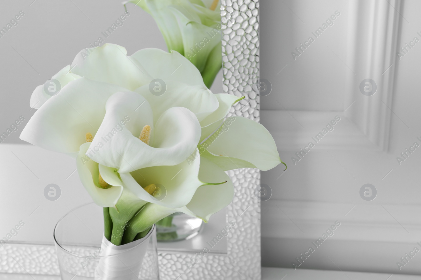 Photo of Beautiful calla lily flowers tied with ribbon in glass vase near mirror, closeup. Space for text