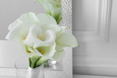 Beautiful calla lily flowers tied with ribbon in glass vase near mirror, closeup. Space for text