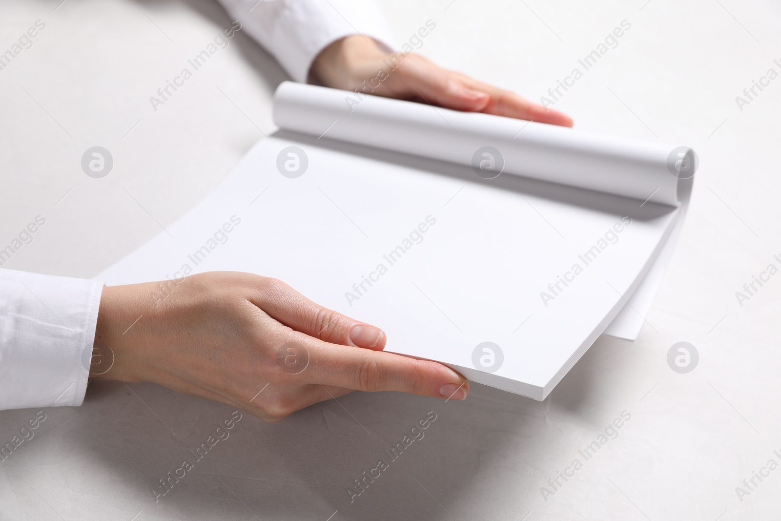 Photo of Woman holding notebook with blank pages at white table, closeup. Mockup for design