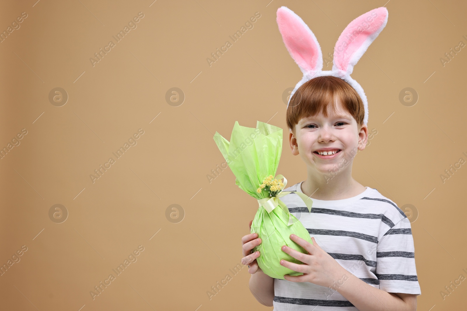 Photo of Easter celebration. Cute little boy with bunny ears and wrapped egg on dark beige background. Space for text