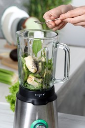 Photo of Woman adding chia seeds into blender with ingredients for green smoothie at white table, closeup
