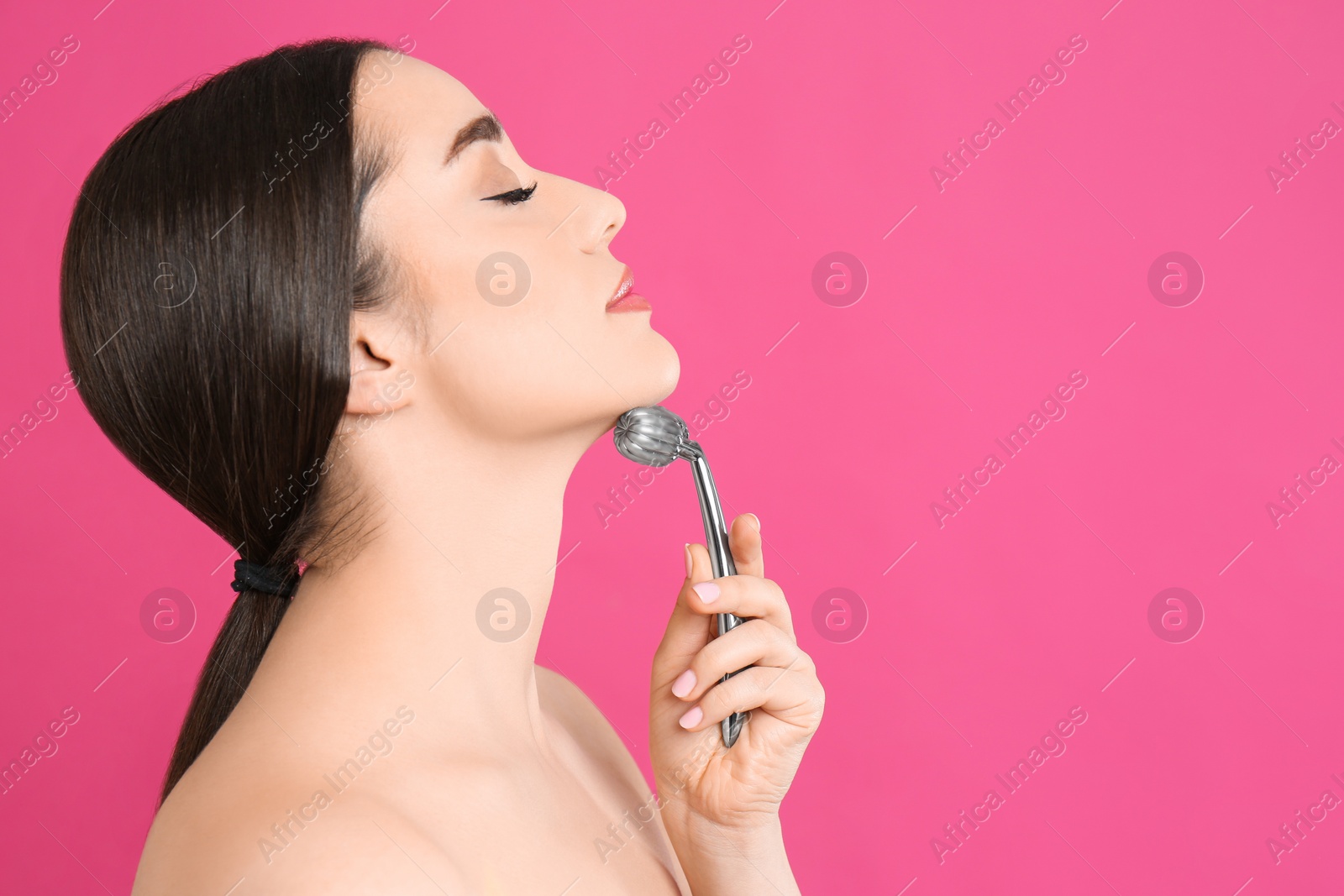 Photo of Woman using metal face roller on pink background, space for text