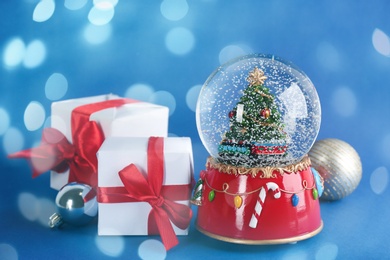 Image of Beautiful Christmas snow globe, gift boxes and ornaments on blue background, bokeh effect