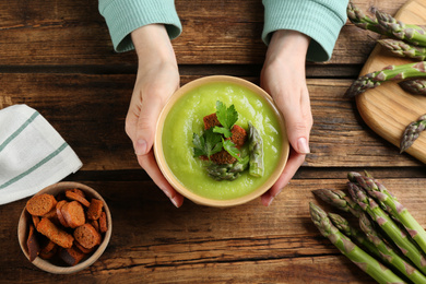 Woman with bowl of asparagus soup at wooden table, top view