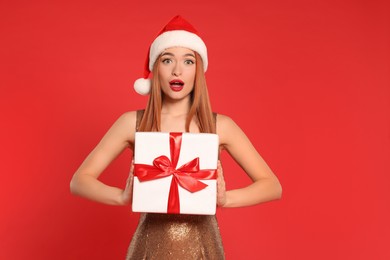 Emotional young woman in Santa hat with Christmas gift on red background