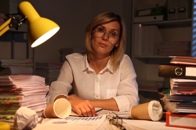 Overwhelmed woman surrounded by documents and paper coffee cups at table in office at night