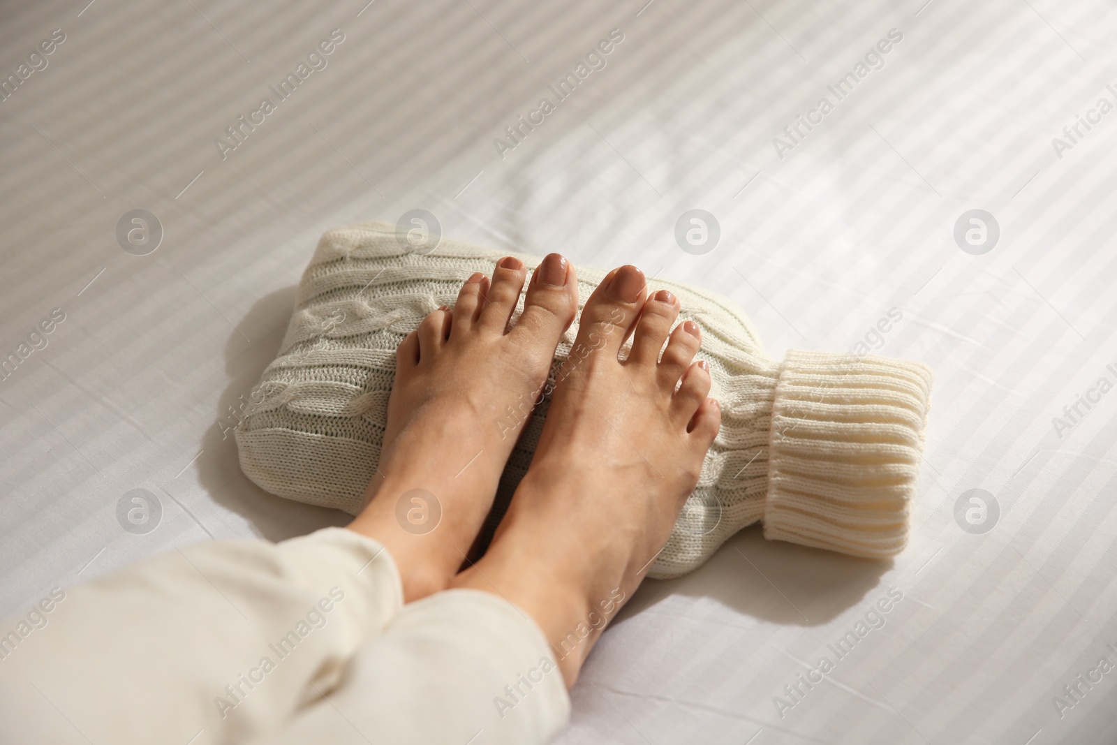 Photo of Woman warming feet with hot water bottle on bed, closeup