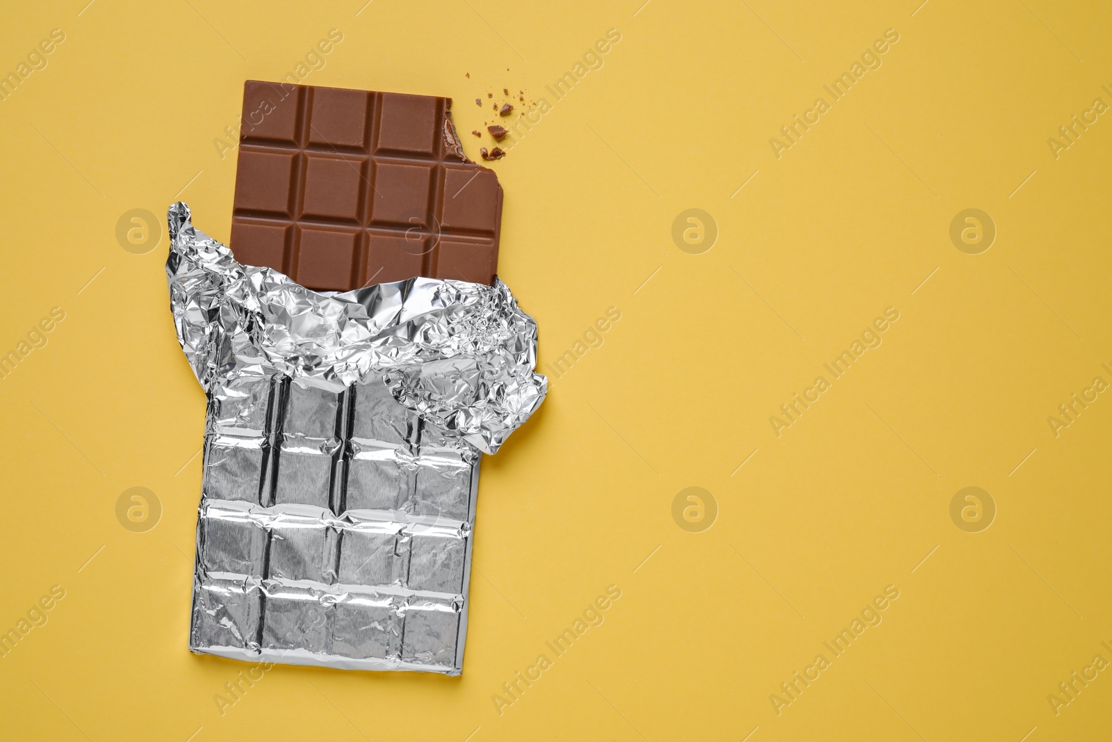 Photo of Bitten milk chocolate bar wrapped in foil on pale yellow background, top view. Space for text