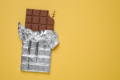 Photo of Bitten milk chocolate bar wrapped in foil on pale yellow background, top view. Space for text