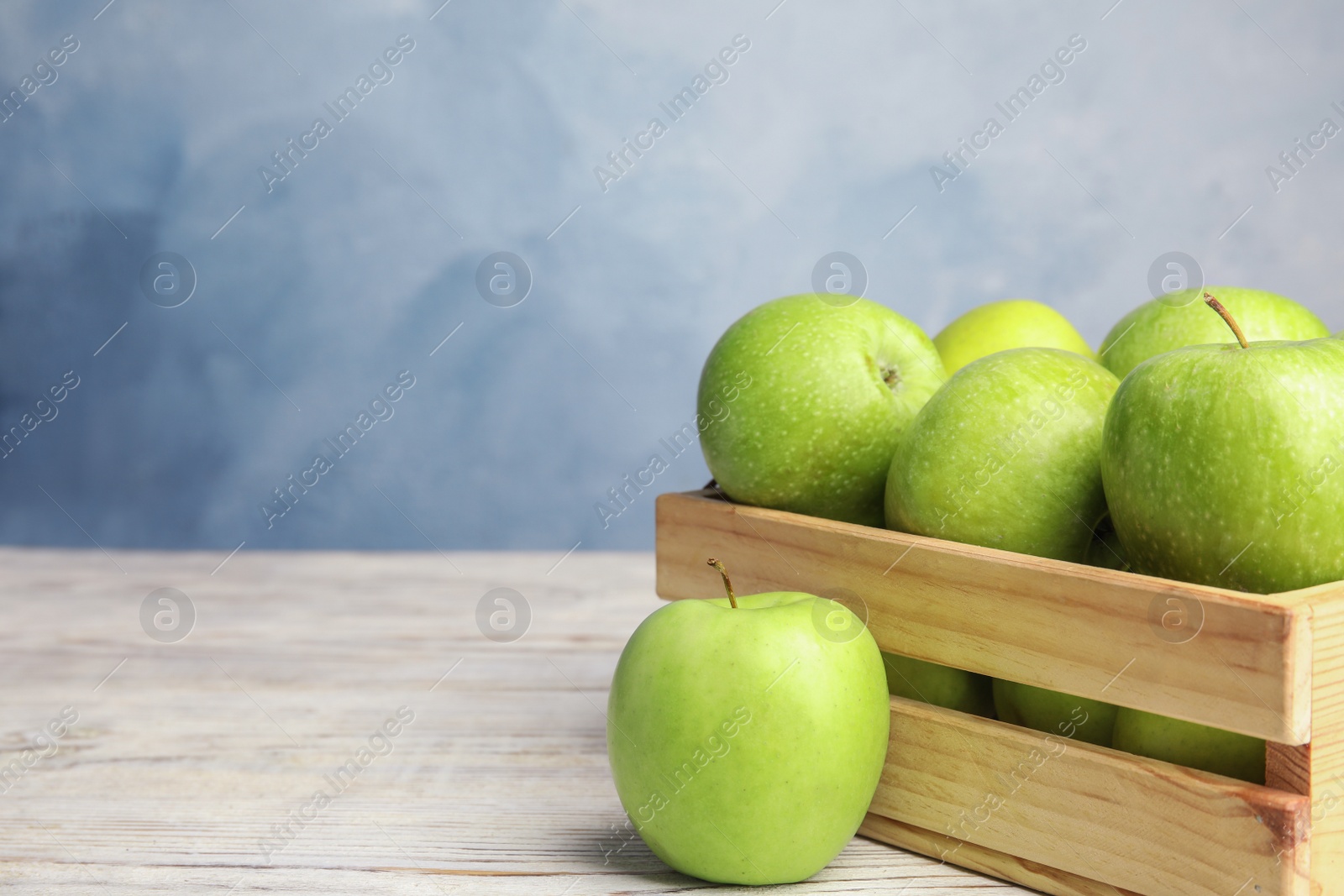 Photo of Wooden crate of fresh ripe green apples on white table against blue background, space for text