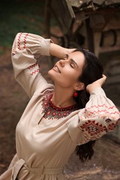 Photo of Beautiful woman wearing embroidered dress and ornate beaded necklace near old wooden well in countryside. Ukrainian national clothes
