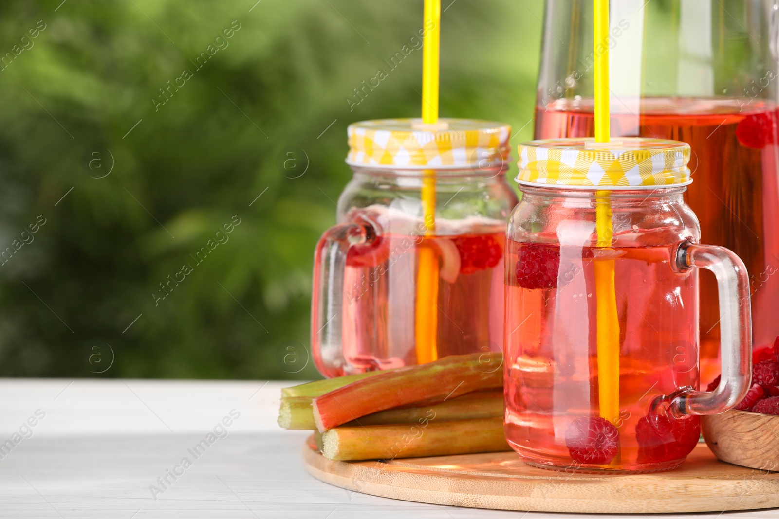 Photo of Tasty rhubarb cocktail with raspberry and stalks on white wooden table outdoors, space for text