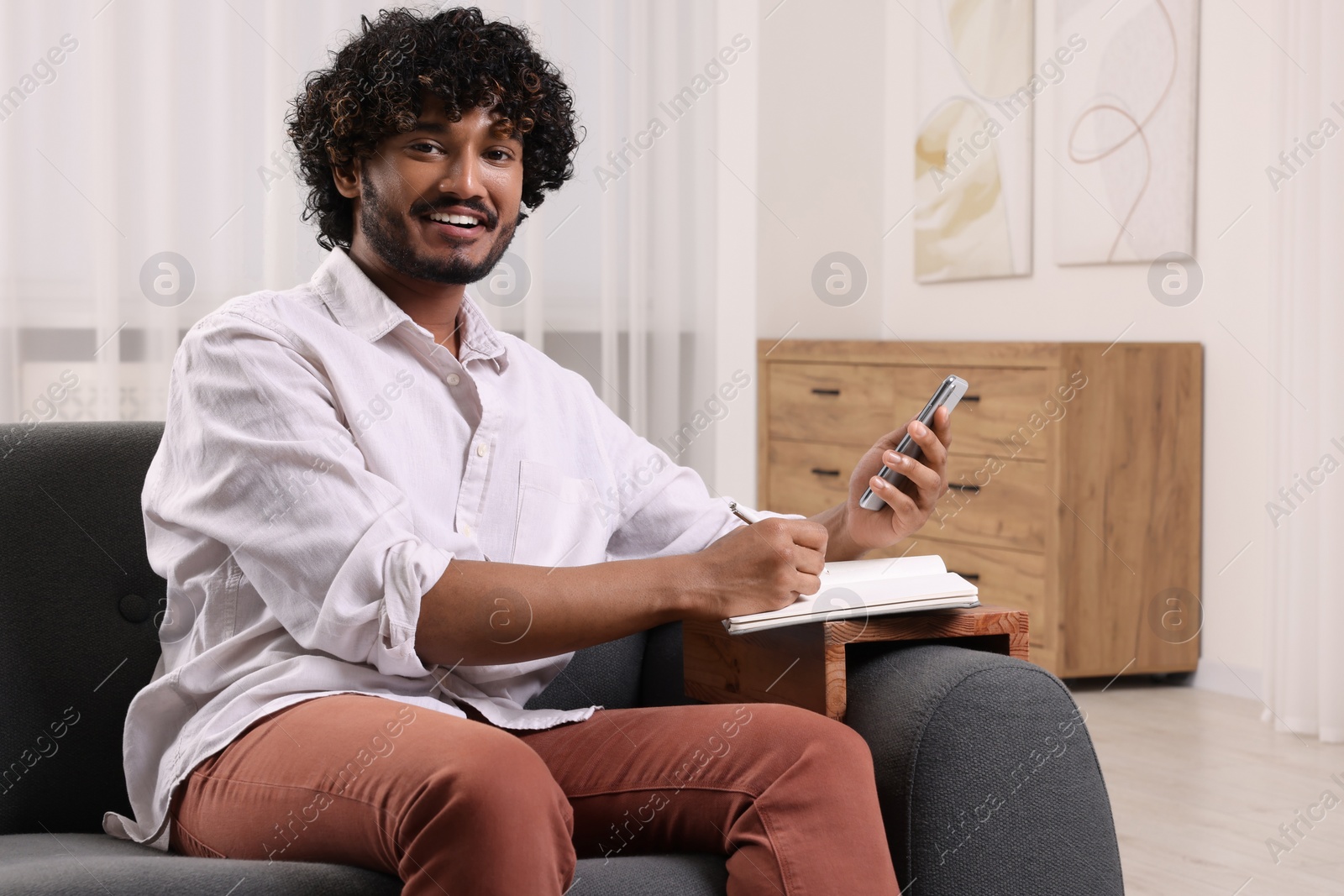 Photo of Happy man using smartphone while writing in notebook on sofa armrest wooden table at home