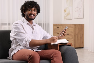Happy man using smartphone while writing in notebook on sofa armrest wooden table at home