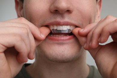 Young man applying whitening strip on his teeth against light grey background, closeup