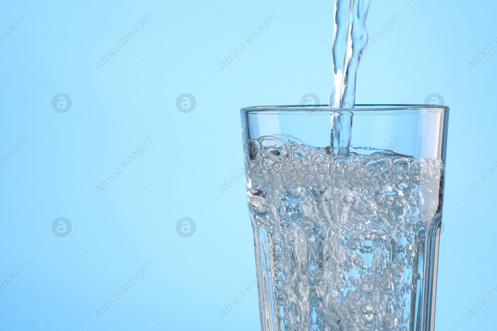 Photo of Pouring water into glass on light blue background, closeup. Space for text