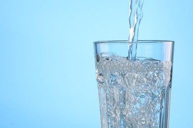 Pouring water into glass on light blue background, closeup. Space for text