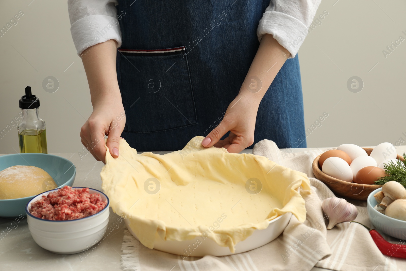 Photo of Woman putting dough for meat pie into baking dish at light grey table, closeup