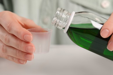 Photo of Woman pouring syrup from bottle into measuring cup at table, closeup. Cold medicine