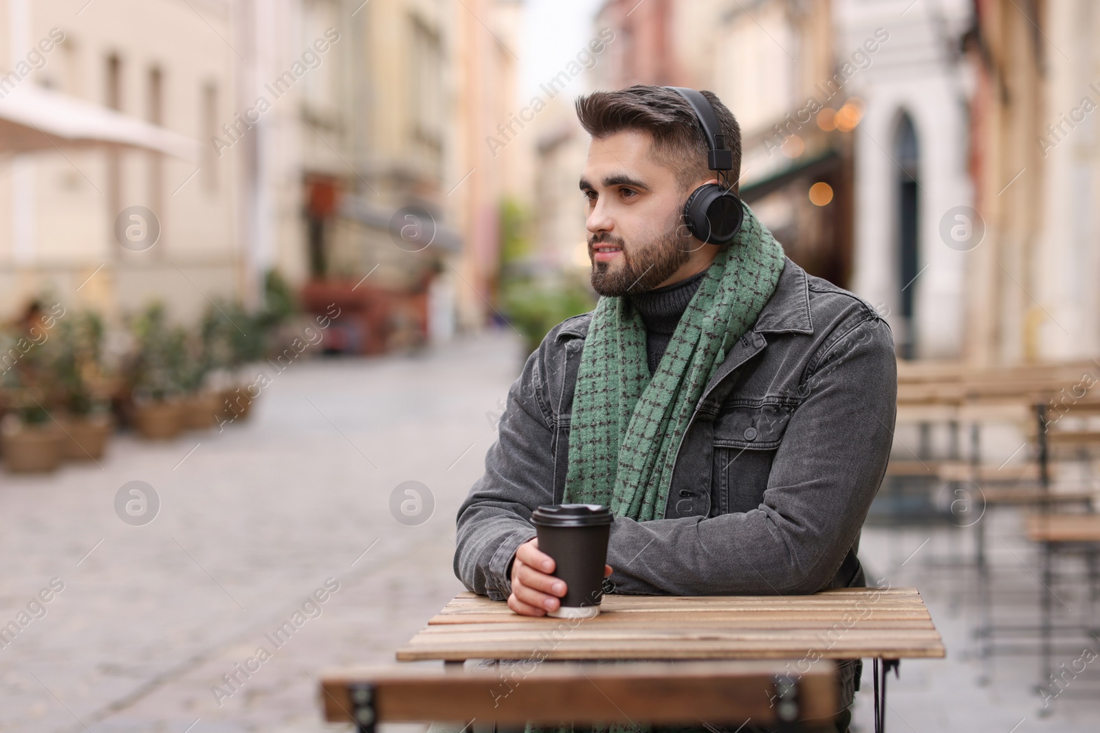 Photo of Handsome man in warm scarf with paper cup listening to music in outdoor cafe. Space for text