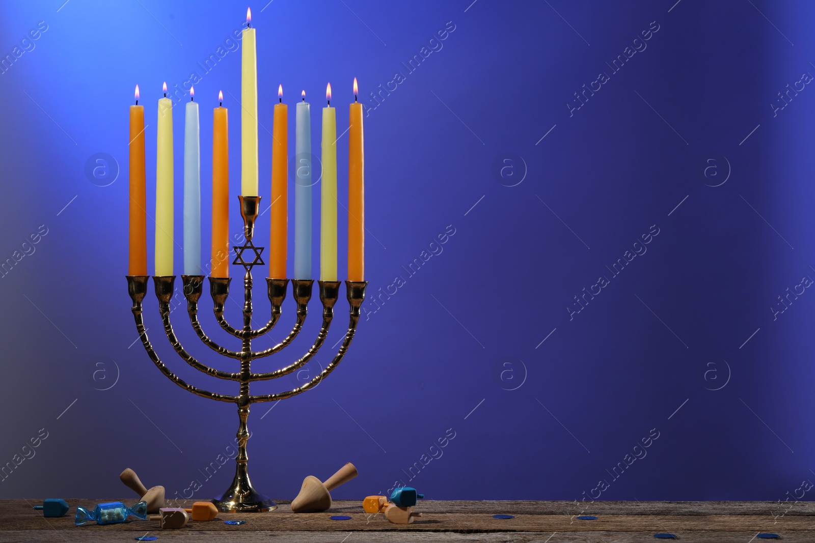 Photo of Hanukkah celebration. Menorah with burning candles and dreidels on wooden table against blue background, space for text