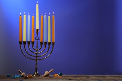 Photo of Hanukkah celebration. Menorah with burning candles and dreidels on wooden table against blue background, space for text