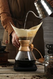 Photo of Woman pouring hot water into glass chemex coffeemaker with paper filter at wooden table, closeup