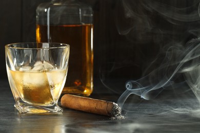 Photo of Bottle, glass of whiskey with ice cubes and smoldering cigar on black wooden table. Space for text