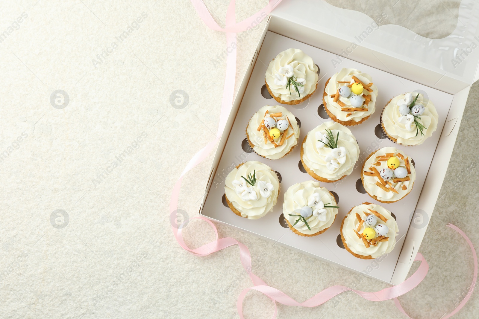 Photo of Tasty Easter cupcakes with vanilla cream in box and pink ribbon on beige table, flat lay. Space for text