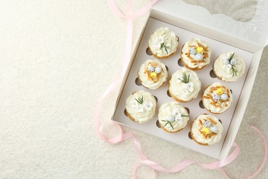 Tasty Easter cupcakes with vanilla cream in box and pink ribbon on beige table, flat lay. Space for text