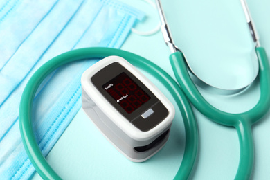 Photo of Modern fingertip pulse oximeter, mask and stethoscope on light blue background, closeup