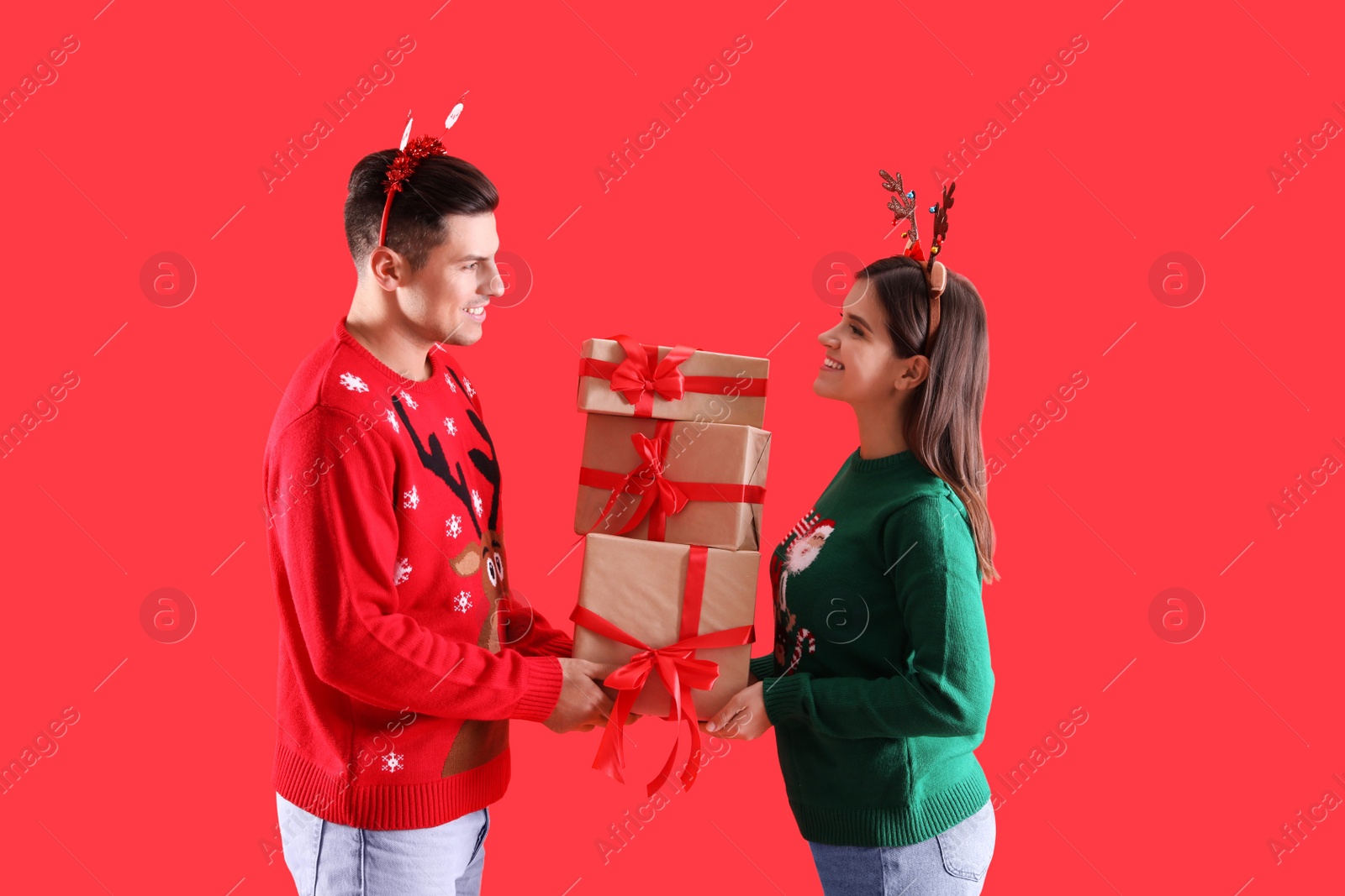 Photo of Beautiful happy couple in Christmas headbands and sweaters holding gifts on red background