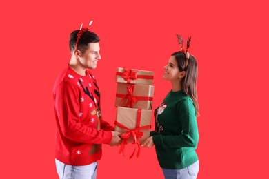 Photo of Beautiful happy couple in Christmas headbands and sweaters holding gifts on red background