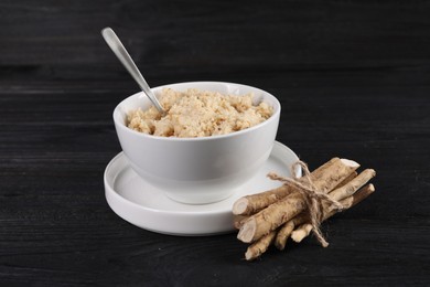 Photo of Bowl of tasty prepared horseradish, spoon and roots on black wooden table