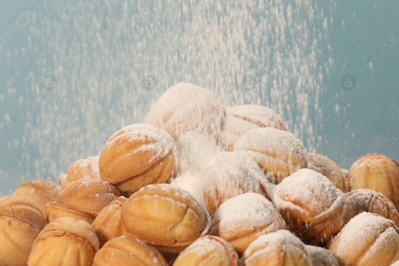 Photo of Sprinkling powdered sugar onto freshly baked walnut shaped cookies against light blue background, closeup. Homemade pastry filled with caramelized condensed milk