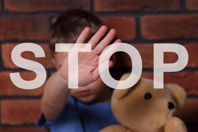 Image of No child abuse. Boy with teddy bear making stop gesture near brick wall, selective focus