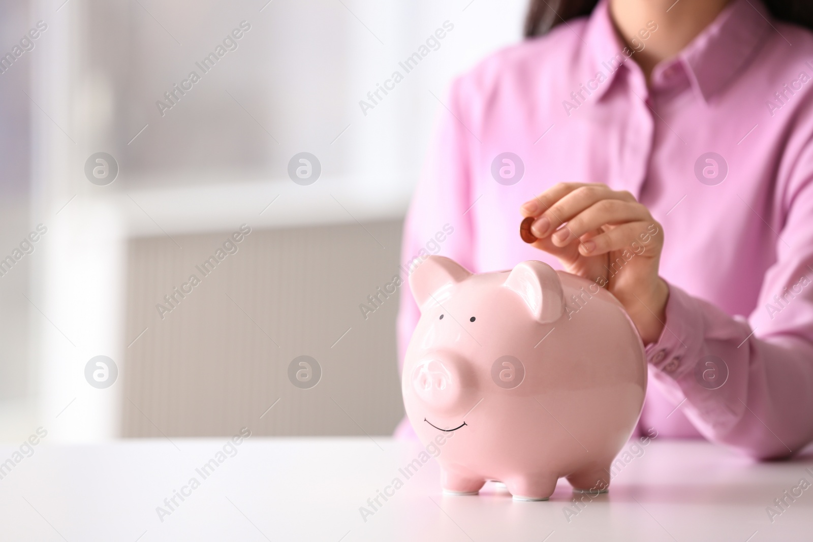 Photo of Woman putting coin into piggy bank at table indoors, closeup. Space for text