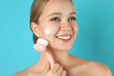 Photo of Young woman washing face with brush and cleansing foam on light blue background. Cosmetic products