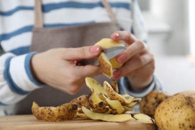 Photo of Woman peeling fresh potato with knife at table, focus on peels