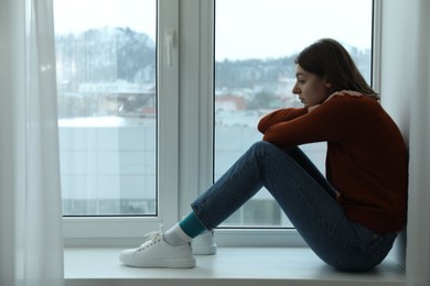 Sad young woman sitting on windowsill near window at home, space for text