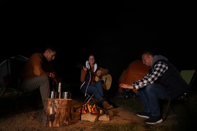 Photo of Group of friends with guitar near bonfire and camping tent outdoors at night