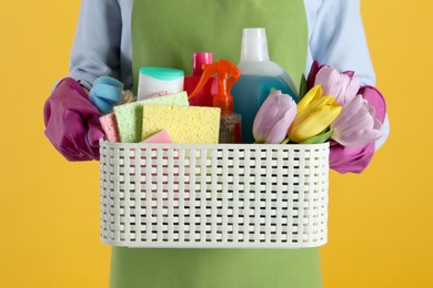 Spring cleaning. Woman holding basket with detergents, flowers and tools on orange background, closeup