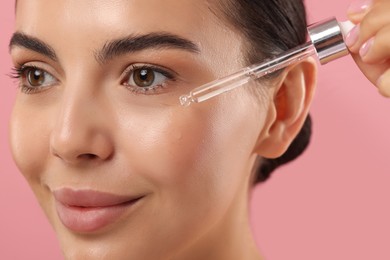 Beautiful young woman applying serum onto her face on pink background, closeup