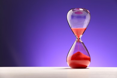 Photo of Hourglass with red flowing sand on white table against purple background. Space for text
