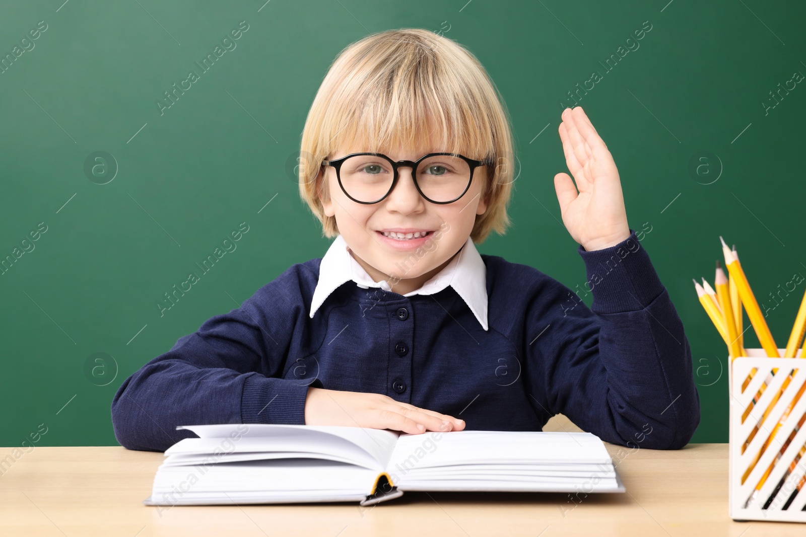 Photo of Happy little school child raising hand while sitting at desk with books near chalkboard