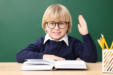Happy little school child raising hand while sitting at desk with books near chalkboard