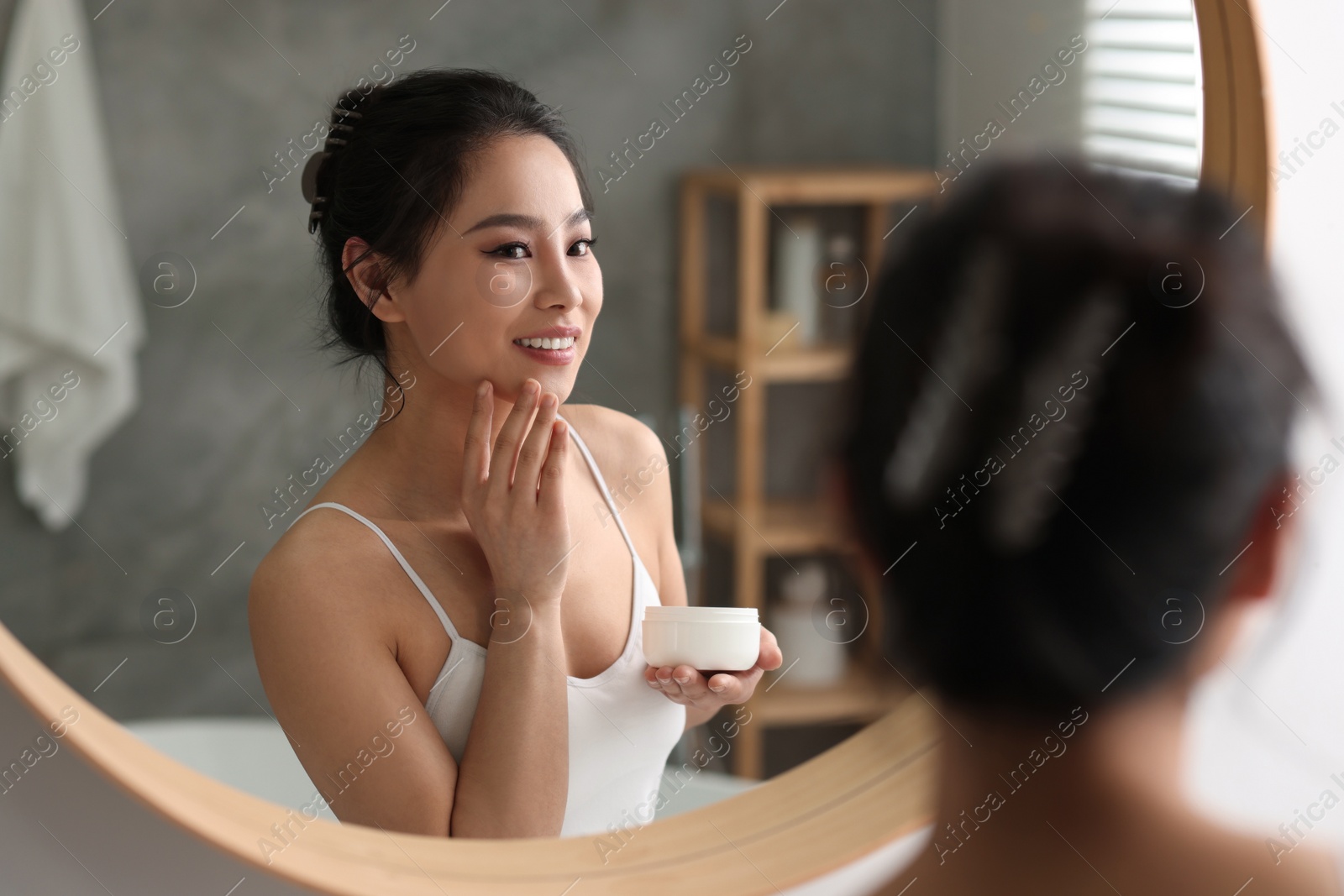 Photo of Happy woman applying face cream near mirror at home